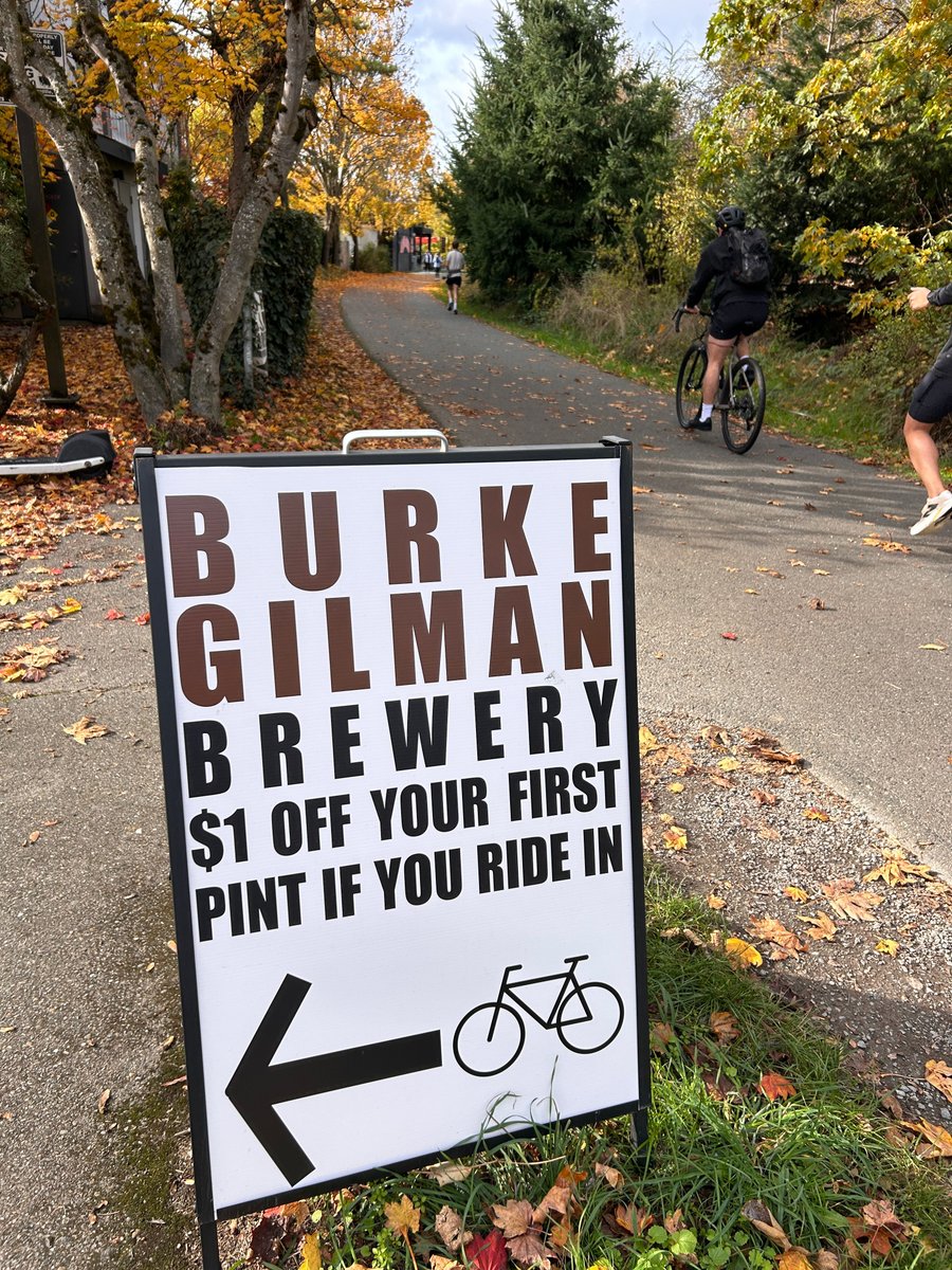 The "$1 off your first pint if you ride in" sign on the Burke-Gilman Trail in fall, with a cyclist riding past